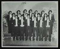 Unidentified group of nineteen escorts in tuxedos in three rows in front of stage with Links sign hanging at back