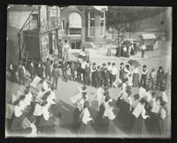 Parade of women with flags in front of prop buildings (Semi-Centennial Parade)
